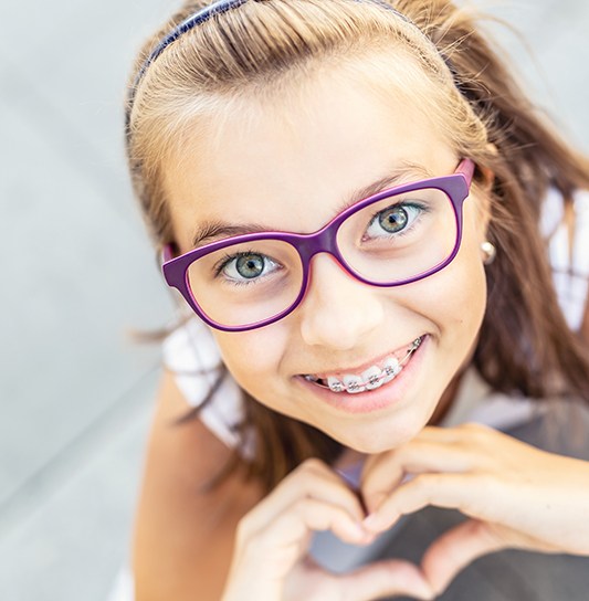 Girl in purple glasses smiling with braces looking up and making a heart with her hands