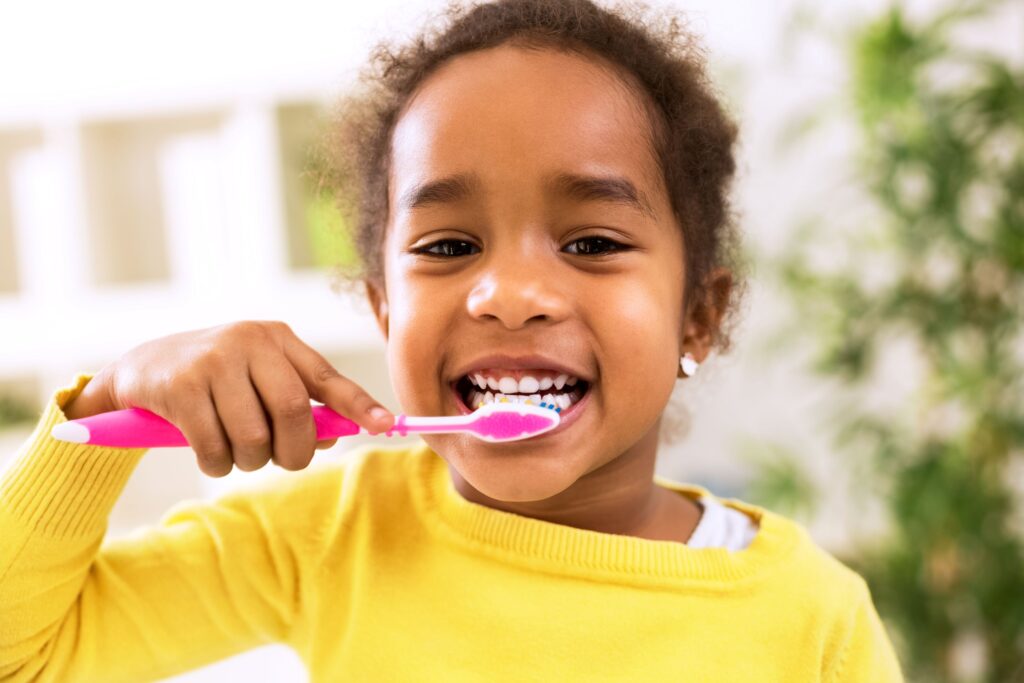 Little girl in yellow sweater brushing teeth with pink and white toothbrush